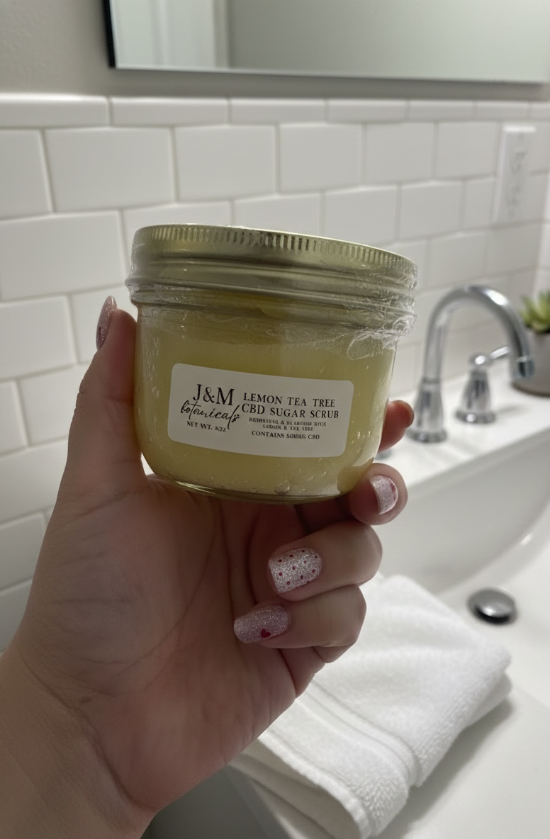 Hand holding a jar of Lemon Tea Tree CBD Sugar Scrub in a bathroom setting.