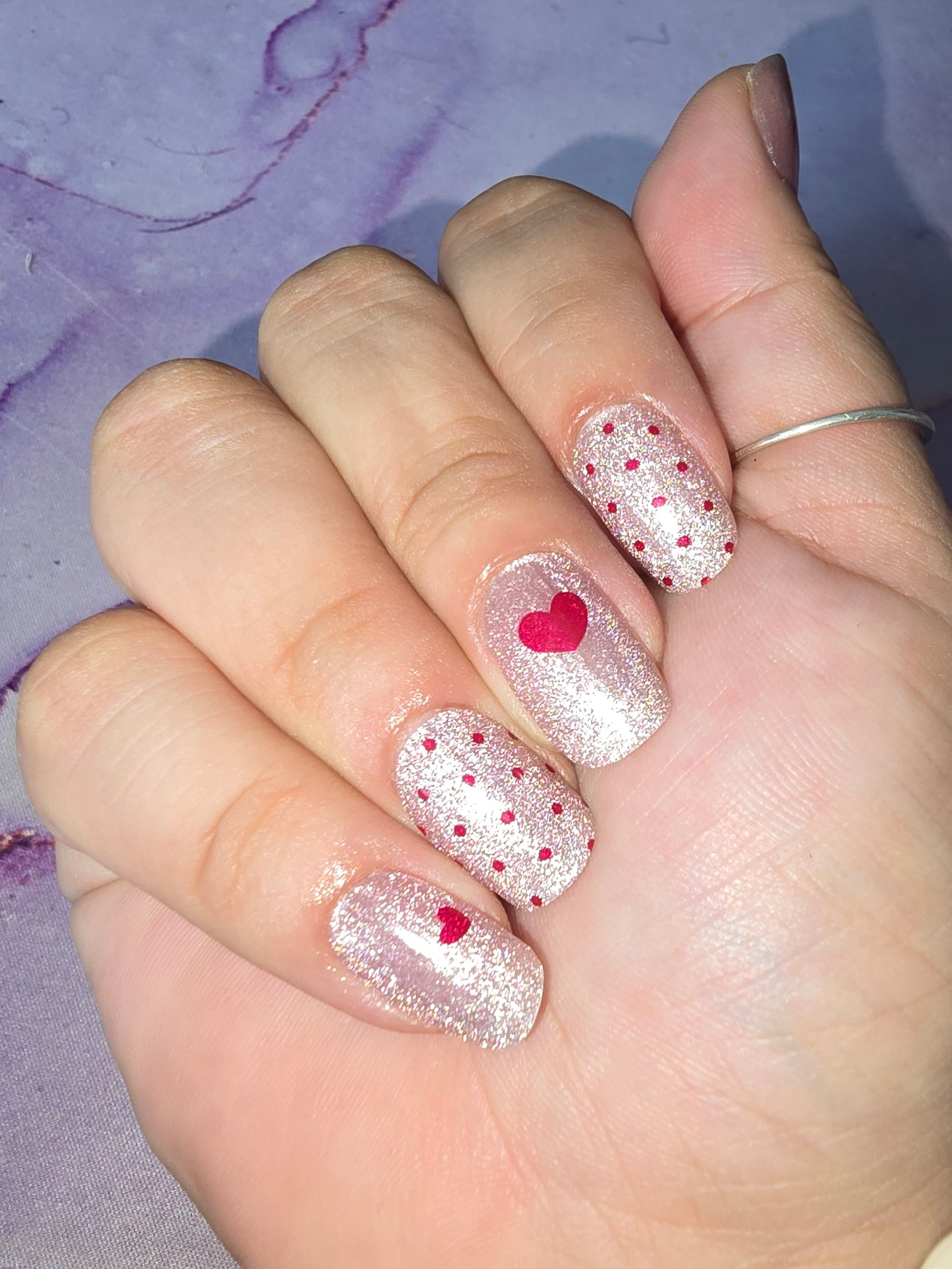 A glittery/shimmer pale pink cat eye base with red polka dots on some nails and red hearts on others.
