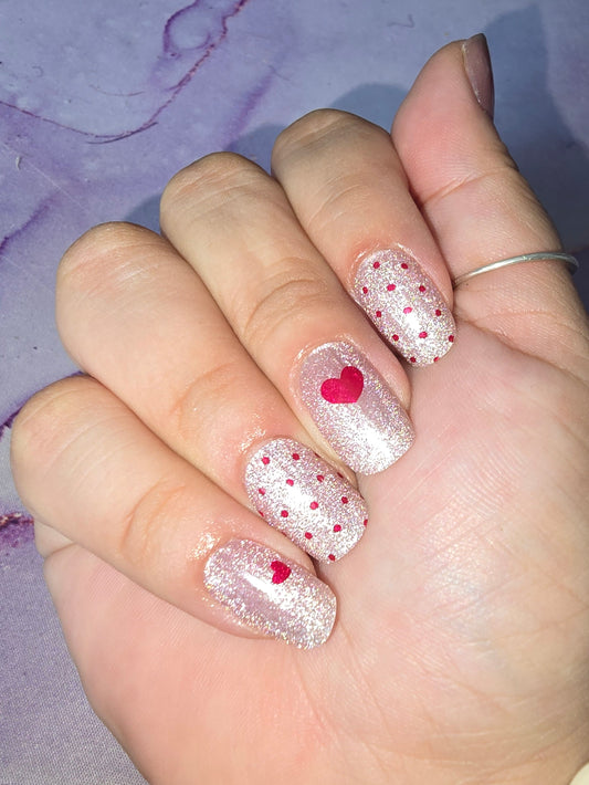 A glittery/shimmer pale pink cat eye base with red polka dots on some nails and red hearts on others.